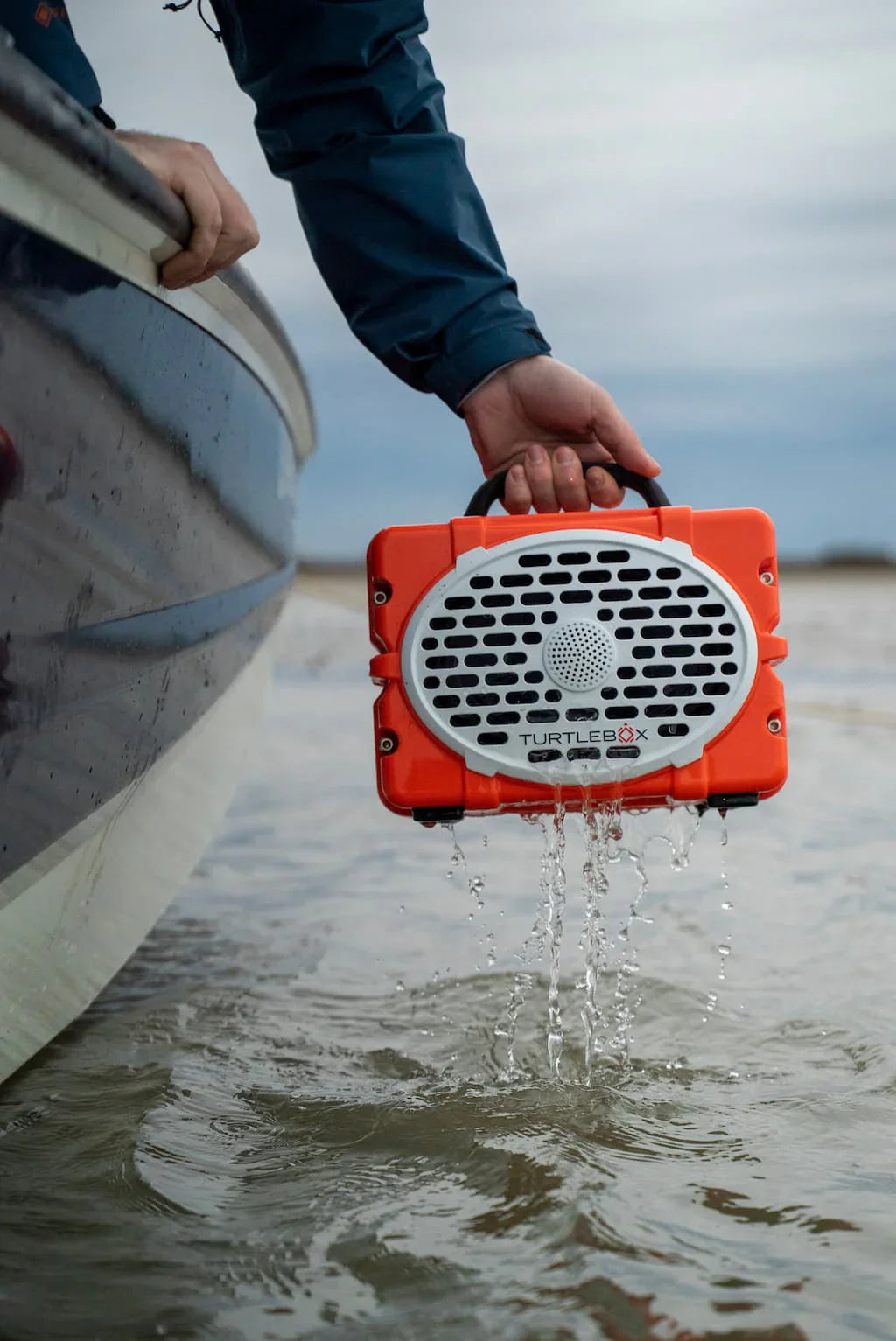 Turtlebox Original Gen3 Speaker in orange being held above water near a boat, designed for outdoor use.